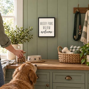 May include: A square wall sign with a black frame and white background featuring the black text MUDDY PAWS WARM welcome. The sign is mounted on a sage green paneled wall above a wooden counter with a pet bowl and a jar of treats.