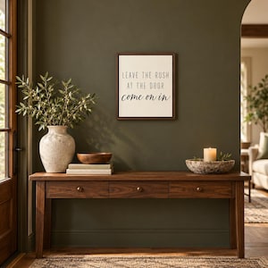 May include: A rectangular wall sign with a dark wood frame and white background features the black text: LEAVE THE RUSH AT THE DOOR come on in. The sign is mounted on a dark olive green wall above a wooden console table with three drawers.