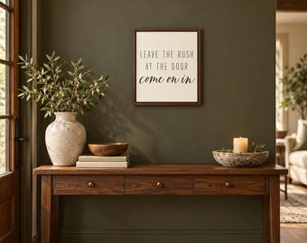 May include: A rectangular wall sign with a dark wood frame and white background features the black text: LEAVE THE RUSH AT THE DOOR come on in. The sign is mounted on a dark olive green wall above a wooden console table with three drawers.