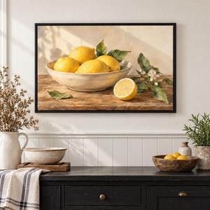 May include: A framed wall art print depicting a still life scene of bright yellow lemons in a ceramic bowl on a wooden surface. One halved lemon and green leaves with a small white flower are placed beside the bowl. The background is a textured, neutral-toned wall with soft shadows cast across the scene.