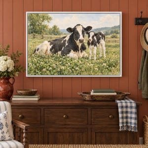 May include: A framed wall art print featuring a black and white cow lying down in a green clover field next to a standing calf. The background shows a rural landscape with trees, a wooden fence, and rolling hills under a cloudy sky.