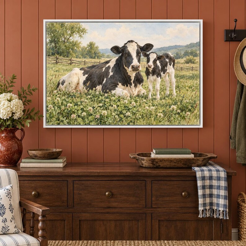 May include: A framed wall art print featuring a black and white cow lying down in a green clover field next to a standing calf. The background shows a rural landscape with trees, a wooden fence, and rolling hills under a cloudy sky.
