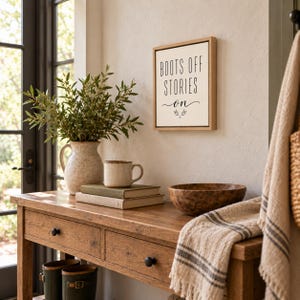 May include: A square wooden-framed wall sign with a cream background and black text that reads BOOTS OFF STORIES ON with a small decorative branch icon. The sign is mounted on a light-colored wall above a wooden console table with drawers.