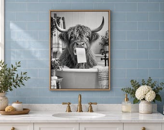 May include: A framed black and white wall art print featuring a shaggy Highland cow sitting in a bathtub and holding a roll of toilet paper in its mouth. The artwork is displayed on a light blue tiled wall above a bathroom vanity sink.