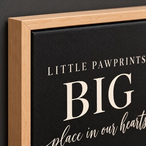 May include: A black canvas wall art piece framed in light-colored wood. The text on the canvas reads LITTLE PAWPRINTS BIG place in our hearts in a mix of serif and script fonts in a light cream color against the textured black background.