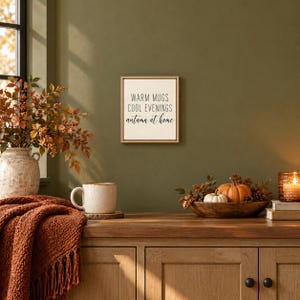 Puede incluir: Un letrero de pared cuadrado con fondo claro y marco de madera fina, que presenta el texto negro WARM MUGS COOL EVENINGS autumn at home. El letrero se muestra en una pared verde oliva sobre una superficie de madera con una taza y decoración.