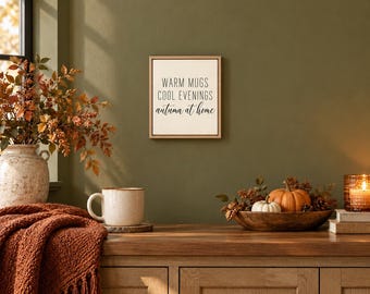 May include: A square wall sign with a light background and a thin wooden frame, featuring the black text WARM MUGS COOL EVENINGS autumn at home. The sign is displayed on an olive green wall above a wooden surface with a mug and decor.