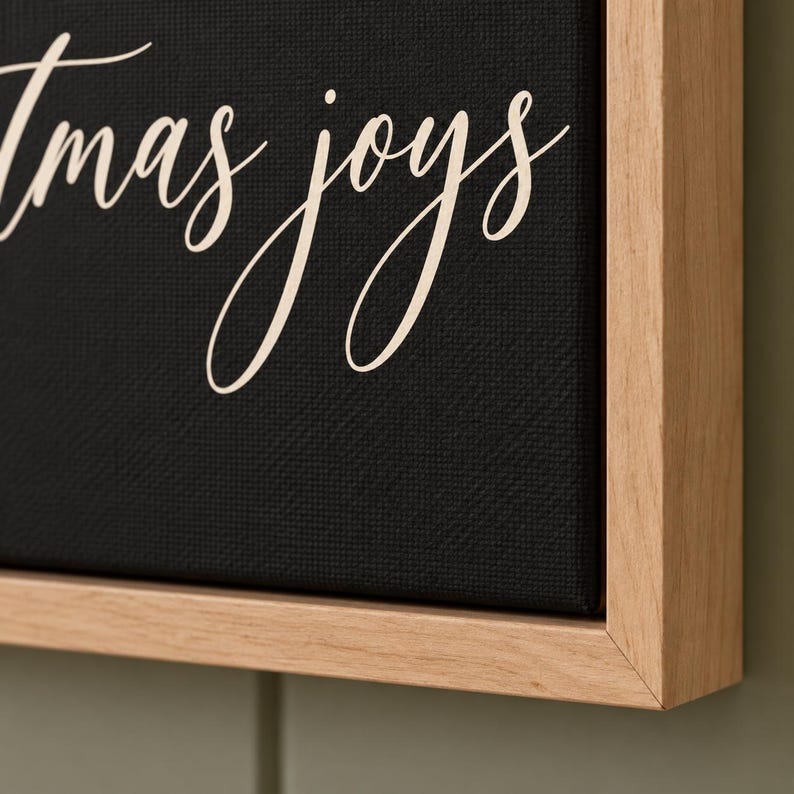May include: A black canvas wall art piece featuring the white cursive text "tmas joys" framed in a light-colored wooden border. The canvas has a visible textured surface, and the lettering is written in a flowing, elegant script font.