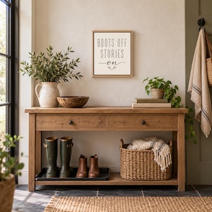 May include: A rectangular wall sign with a light background and a thin wooden frame, featuring the black text "BOOTS OFF STORIES ON" with a small decorative branch icon. The sign is displayed above a wooden console table with two drawers.