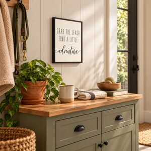 Op de afbeelding: Een rechthoekig wandbord met een zwarte lijst met de tekst "GRAB THE LEASH FIND A LITTLE adventure" in zwarte letters op een witte achtergrond, gemonteerd op een witte paneelwand boven een groene houten kast met een houten blad.