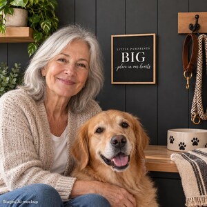 May include: A square black wooden sign with a light wood frame featuring the white text LITTLE PAWPRINTS BIG place in our hearts. The sign is mounted on a dark paneled wall next to a brown leather leash and a white ceramic pet bowl.