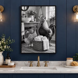 Op de afbeelding: Een rechthoekige wandkunstprint met zwarte lijst, voorzien van een zwart-witfoto van een haan en een kip naast een gevlochten mand vol witte handdoeken, geplaatst in een badkameromgeving met een wastafel en meubel.