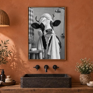 May include: A framed black and white wall art print featuring a cow wearing a towel on its head and wrapped in a towel, holding a bath brush in a tiled shower setting. The artwork is displayed on a terracotta-colored wall above a dark sink.