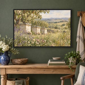 May include: A framed landscape painting depicting three wooden beehives situated in a lush meadow filled with lavender and clover, set against a backdrop of rolling hills, trees, and a soft sky, all enclosed in a simple black frame.