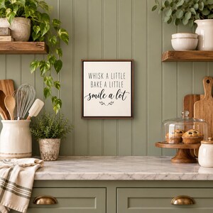 May include: A rectangular wall sign with a dark wood frame featuring the black text "WHISK A LITTLE BAKE A LITTLE smile a lot" centered on a white background, accented by a small decorative leaf illustration at the bottom.