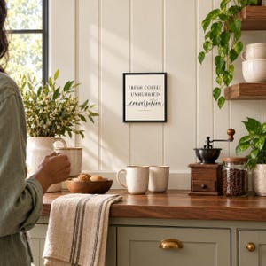 May include: A square wall sign with a black frame and white background featuring the black text FRESH COFFEE UNHURRIED conversation. The sign is displayed in a kitchen setting with coffee mugs, a wooden bowl, and a coffee grinder.
