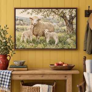 May include: A framed wall art print depicting a large sheep standing in a grassy field with two small lambs under a blooming tree. The scene features rolling hills in the background, soft natural lighting, and a pastoral landscape setting.