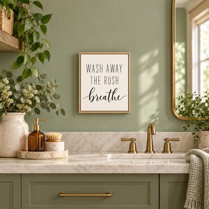May include: A rectangular wall sign with a light wood frame featuring the black text "WASH AWAY THE RUSH breathe" on a white background. The sign is mounted on a sage green wall above a marble countertop with a sink, faucet, and accessories.