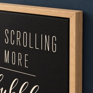 May include: A black canvas wall sign framed in light-colored wood. The sign features white text that reads SCROLLING MORE, with a horizontal line underneath and a partial word in a cursive script font visible at the bottom.
