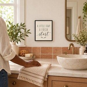 May include: A square wall sign with a black frame and a cream-colored background featuring the black text A LITTLE SOAP A FRESH start with a small decorative leaf illustration centered below the words on the wall.