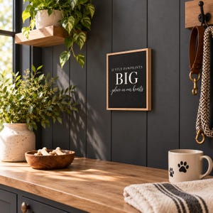 May include: A square wall sign with a light wood frame and black background featuring the white text "LITTLE PAWPRINTS BIG place in our hearts". The sign is mounted on a dark vertical paneled wall near a wooden shelf and a hook.