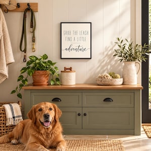 Op de afbeelding: Een rechthoekig wandbord met een witte achtergrond en zwarte tekst die luidt GRAB THE LEASH FIND A LITTLE adventure. Het bord is zwart omlijst en bevestigd aan een witte paneelwand boven een groene houten opbergbank.