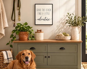 May include: A rectangular wall sign with a white background and black text that reads GRAB THE LEASH FIND A LITTLE adventure. The sign is framed in black and mounted on a white paneled wall above a green wooden storage bench.