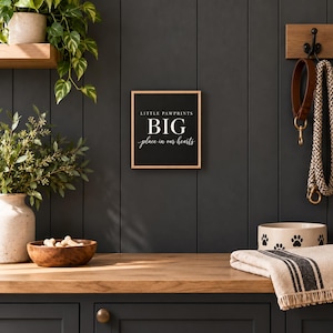 May include: A square wooden sign with a black background and a light wood frame. The text in white reads LITTLE PAWPRINTS BIG place in our hearts. The sign is mounted on a dark gray wall, positioned between a green plant and a pet leash.