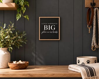 May include: A square wooden sign with a black background and a light wood frame. The text in white reads LITTLE PAWPRINTS BIG place in our hearts. The sign is mounted on a dark gray wall, positioned between a green plant and a pet leash.