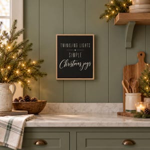 May include: A rectangular wall sign with a light wood frame and a black background. The text in white reads TWINKLING LIGHTS, a small star symbol, SIMPLE, and Christmas joys in a cursive script font. The sign is mounted on a green wall.
