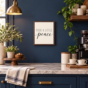 May include: A square wall sign with a dark wood frame and a cream-colored background. The text in black reads "POUR A LITTLE" in a simple sans-serif font, with the word "peace" written below in a flowing, cursive script.