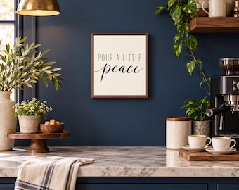 May include: A square wall sign with a dark wood frame and a cream-colored background. The text in black reads "POUR A LITTLE" in a simple sans-serif font, with the word "peace" written below in a flowing, cursive script.