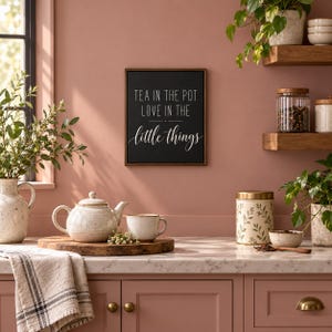 May include: A rectangular black wall sign with white text that reads TEA IN THE POT LOVE IN THE little things, framed in dark wood. The sign is displayed on a dusty rose wall above a kitchen counter with a ceramic teapot and mug.