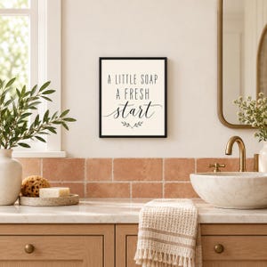 May include: A rectangular wall sign with a black frame and a cream-colored background. The text reads A LITTLE SOAP A FRESH start in black lettering, with a small decorative leaf graphic centered below the text.
