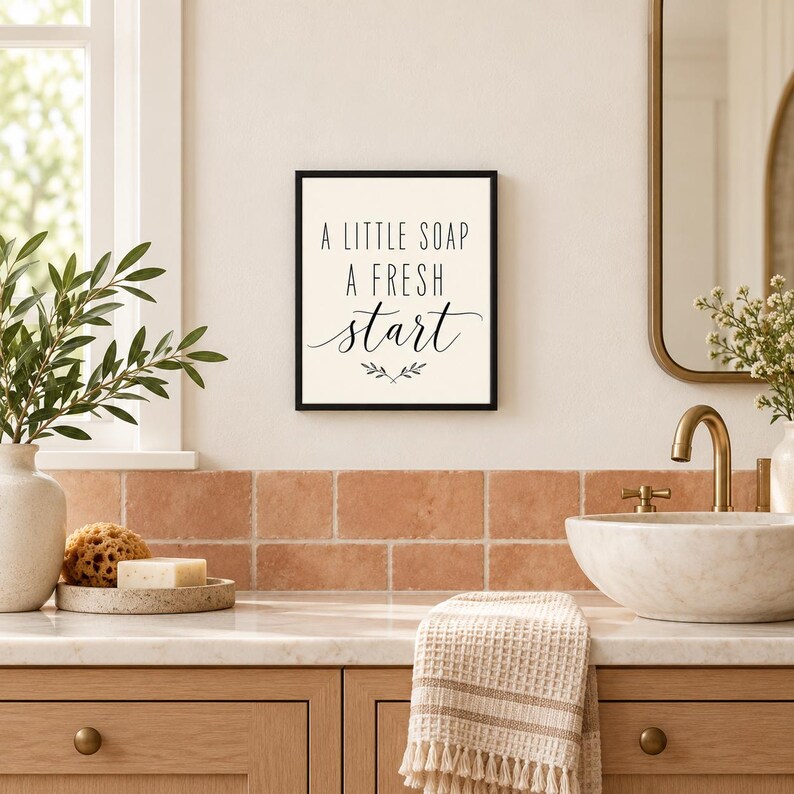 May include: A rectangular wall sign with a black frame and a cream-colored background. The text reads A LITTLE SOAP A FRESH start in black lettering, with a small decorative leaf graphic centered below the text.