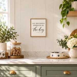 May include: A square wall sign with a light wood frame and a white background featuring the black text "ALL DECISIONS APPROVED BY the dog" in a combination of serif and script fonts, centered on a white paneled wall.