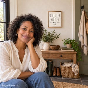 May include: A square wooden wall sign with a light frame featuring the black text BOOTS OFF STORIES ON. The sign is displayed on a wall above a wooden console table holding a ceramic pitcher, a wooden bowl, a potted plant, and books.