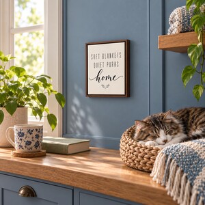 Może przedstawiać: Kwadratowy znak ścienny z ciemną drewnianą ramą i białym tłem, zawierający czarny napis "SOFT BLANKETS QUIET PURRS home" oraz małą dekoracyjną ikonę gałązki wyśrodkowaną pod tekstem na niebieskiej ścianie.