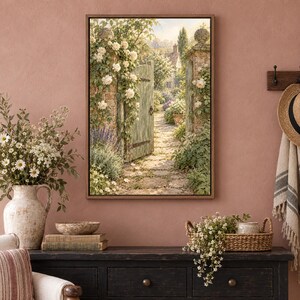 May include: A framed wall art print depicting a stone garden gate opening onto a sunlit cobblestone path, surrounded by climbing white roses and lush greenery. The scene features a rustic wooden gate, stone pillars, and a cottage in the distance.