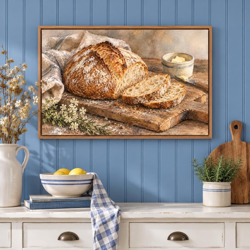 May include: A framed wall art print depicting a rustic loaf of sourdough bread on a wooden cutting board, with two sliced pieces, a small bowl of butter, and a white cloth napkin. The scene is captured in warm, natural lighting tones.