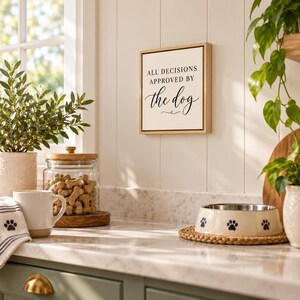 May include: A square wooden framed wall sign with a white background and black text that reads ALL DECISIONS APPROVED BY the dog. The sign features a simple serif and script font combination, mounted on a white paneled wall surface.