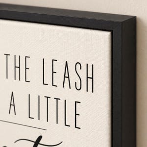 Op de afbeelding: Een ingelijst canvas kunstwerk met een cr&egrave;mekleurige, getextureerde achtergrond en zwarte schreefloze tekst met de woorden THE LEASH A LITTLE. Het kunstwerk is gevat in een dunne, matzwarte lijst met een strak profiel.