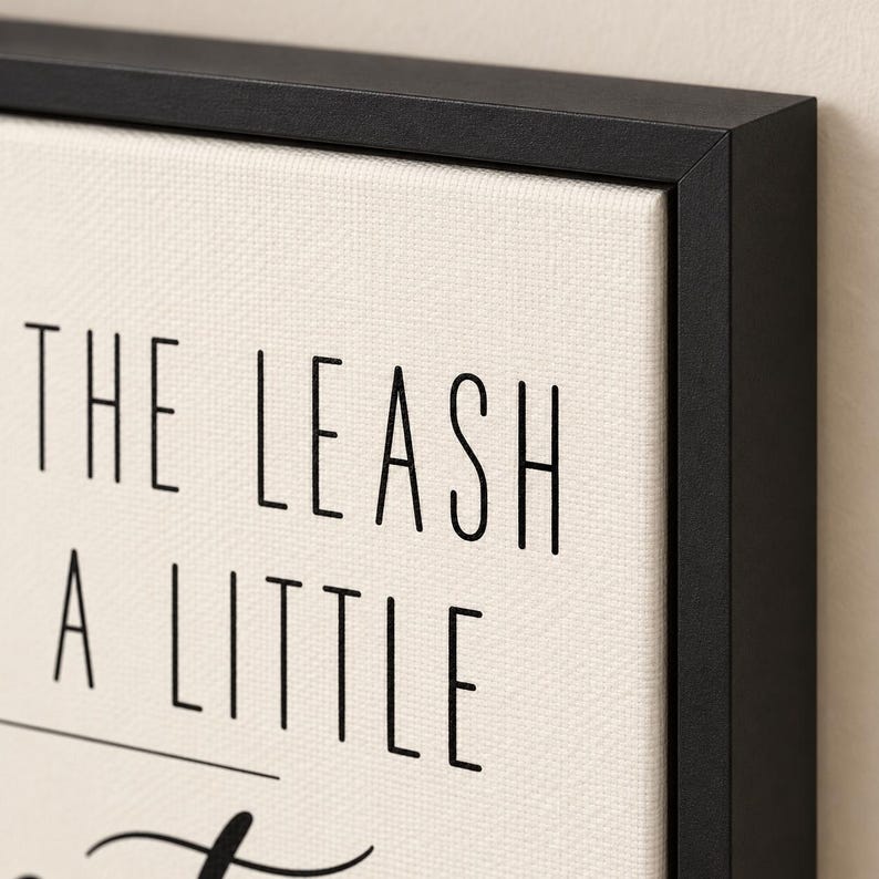 Op de afbeelding: Een ingelijst canvas kunstwerk met een cr&egrave;mekleurige, getextureerde achtergrond en zwarte schreefloze tekst met de woorden THE LEASH A LITTLE. Het kunstwerk is gevat in een dunne, matzwarte lijst met een strak profiel.