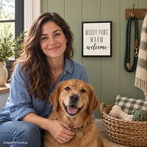 May include: A square wall sign with a black frame and white background featuring the black text MUDDY PAWS WARM welcome. The sign is mounted on a sage green paneled wall next to a dark green dog leash hanging on a wooden hook.