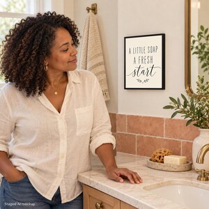 May include: A square wall sign with a black frame and a white background featuring the black text A LITTLE SOAP A FRESH start with a small decorative leaf graphic centered below the text, mounted on a bathroom wall.