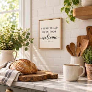 May include: A rectangular wall sign with a light-colored background and a thin wooden frame. The sign features black text that reads FRESH BREAD OPEN DOOR welcome, with the word welcome written in a cursive script font.