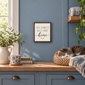 Może przedstawiać: Kwadratowy znak ścienny z ciemnobrązową ramą i kremowym tłem. Tekst brzmi SOFT BLANKETS QUIET PURRS home czarnymi literami, z małym ozdobnym akcentem pod słowem home.