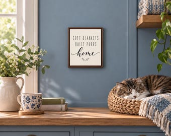 May include: A square wall sign with a dark brown frame and a cream-colored background. The text reads SOFT BLANKETS QUIET PURRS home in black lettering, with a small decorative flourish underneath the word home.