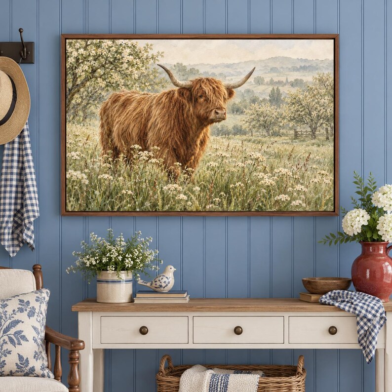 Puede incluir: Una impresi&oacute;n de arte mural rectangular que muestra una vaca Highland de pelo largo de pie en un campo de flores silvestres blancas. La escena incluye colinas y &aacute;rboles al fondo, todo dentro de un marco de madera marr&oacute;n.