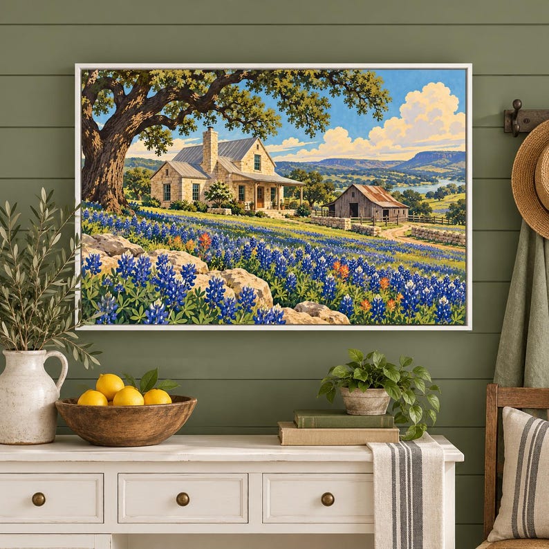 May include: A framed landscape painting depicts a stone house and a rustic barn nestled in a field of bluebonnets under a bright sky. The scene features a large tree, rolling hills, and a distant river, all rendered in vibrant colors.